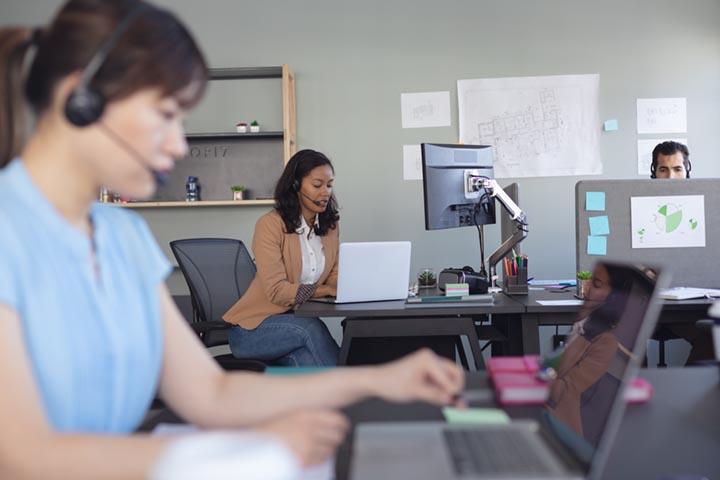 A modern office setting with employees at their desks, including a woman wearing a headset and others working on laptops.