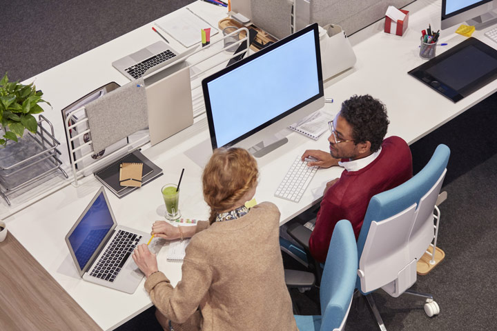 Two people working together at a desk with a desktop computer and laptop, discussing notes and digital content.