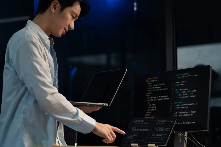 A developer standing and pointing at code on a monitor while holding a laptop in a dimly lit workspace.