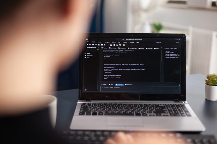 A close-up view of a laptop screen displaying programming code, with a person typing in the foreground.