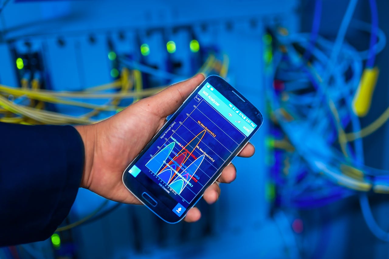 Hand holding a smartphone displaying a network performance graph, with network cables and servers in the background.
